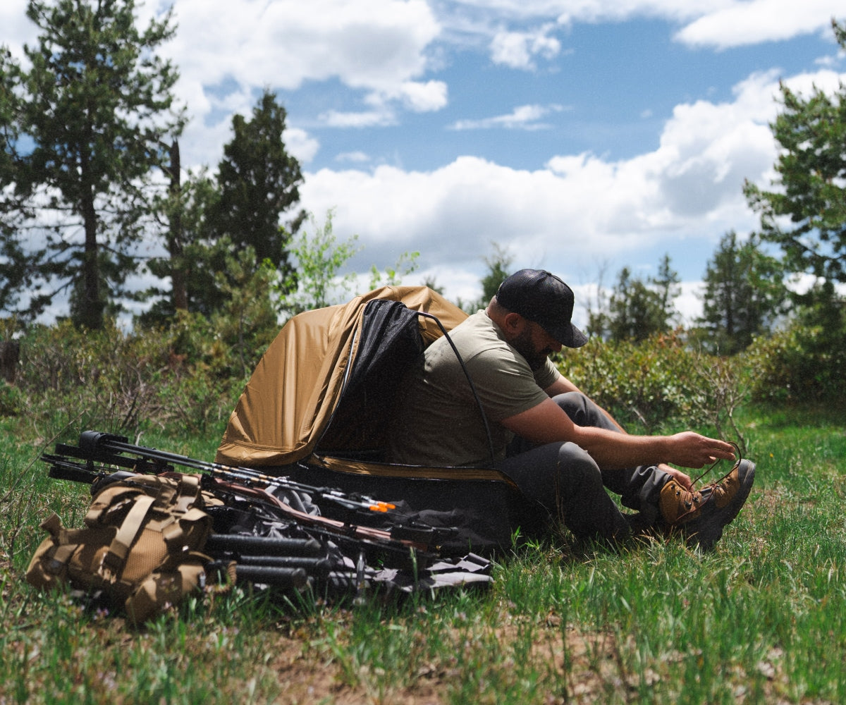 Man sitting outdoors with hunting gear and a waterproof canvas bedroll structure.